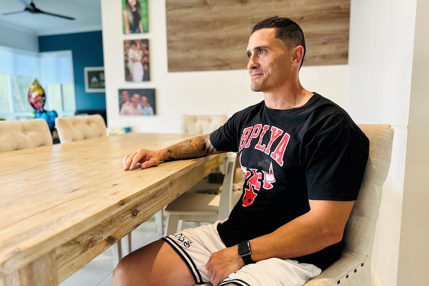 A man wearing a black t shirt sitting at a kitchen table and looking into the distance.