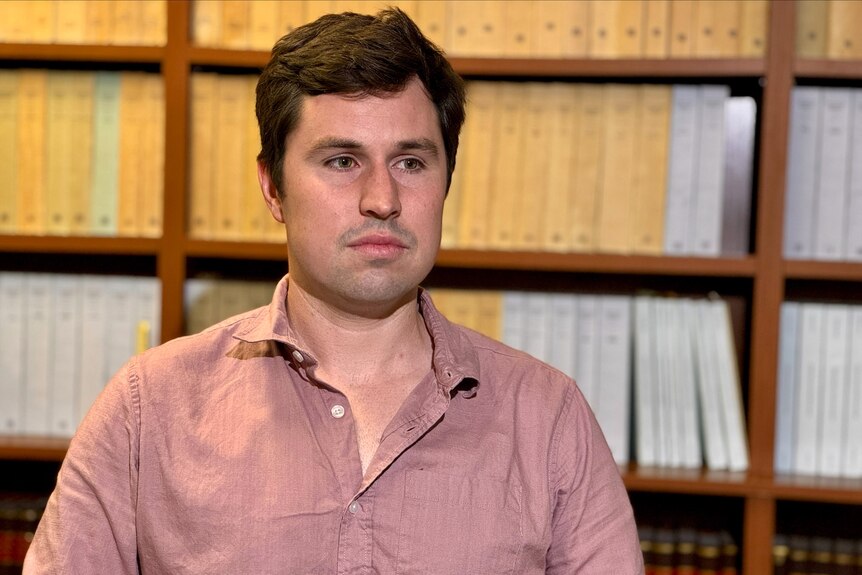 A man wearing a light maroon shirt being interviewed sitting in front of a book case.
