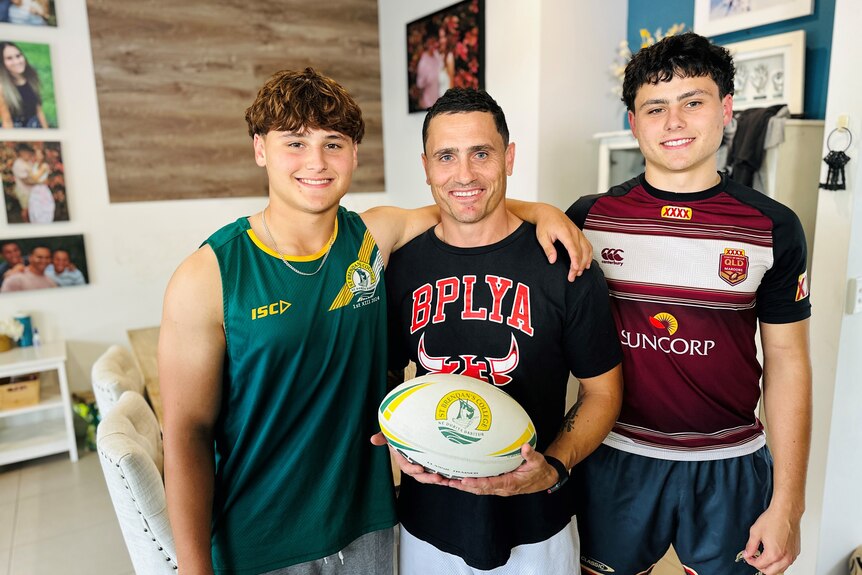 A father standing between two sons in a living room surrounded by family photos.