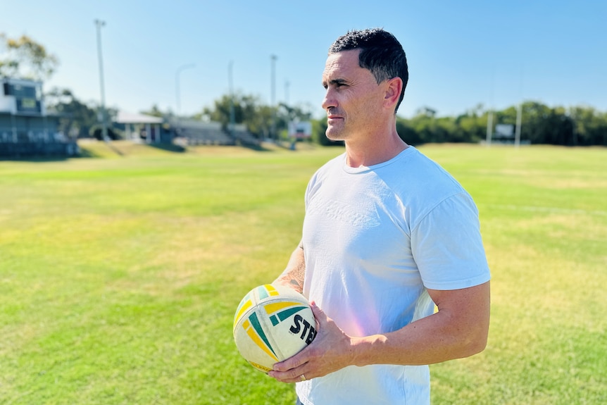 A man wearing a white t shirt and holding a football standing on a footy field.