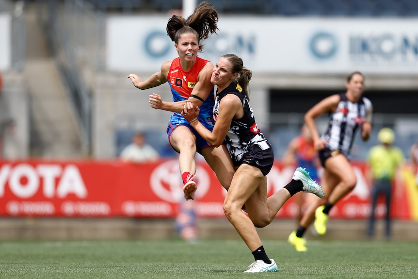 A Melbourne Demons AFLW player kicks the ball in mid-air as she is tackled by a Collingwood defender.