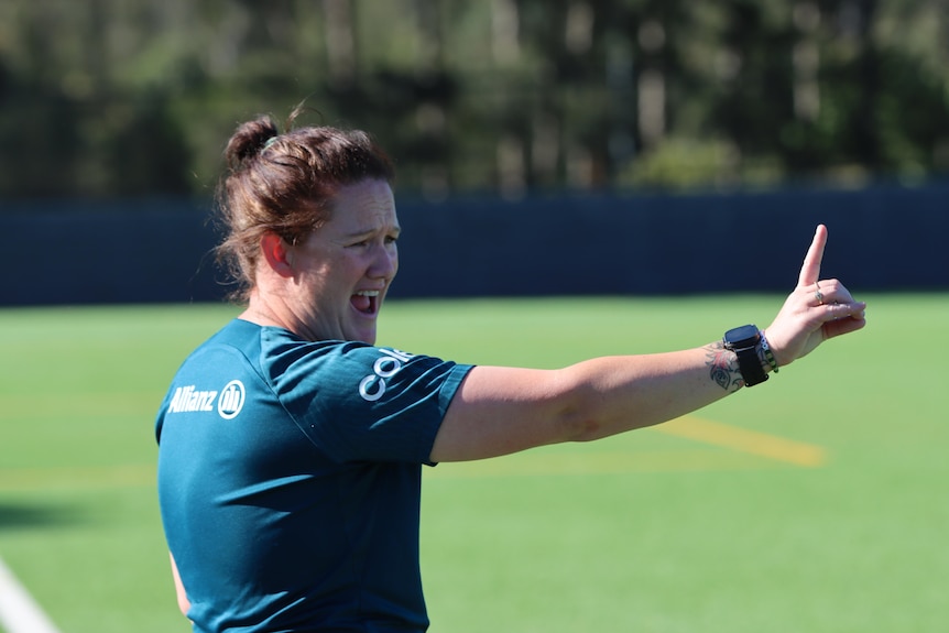 A woman with her hair in a bun and wearing a dark green training jersey is on the sidelines of a soccer field, hand outstretched