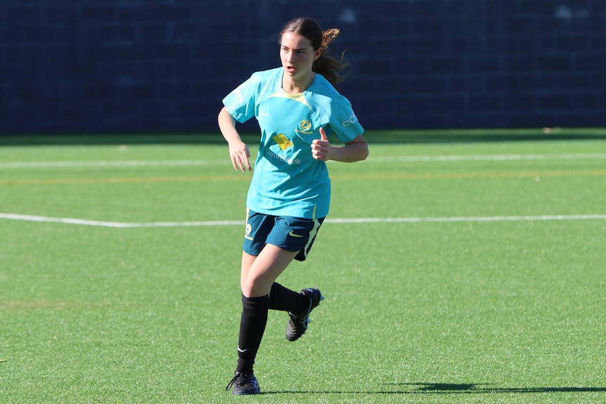 A woman wearing a turquoise soccer jersey, green shorts, black socks and shoes runs on a backdrop of green grass.