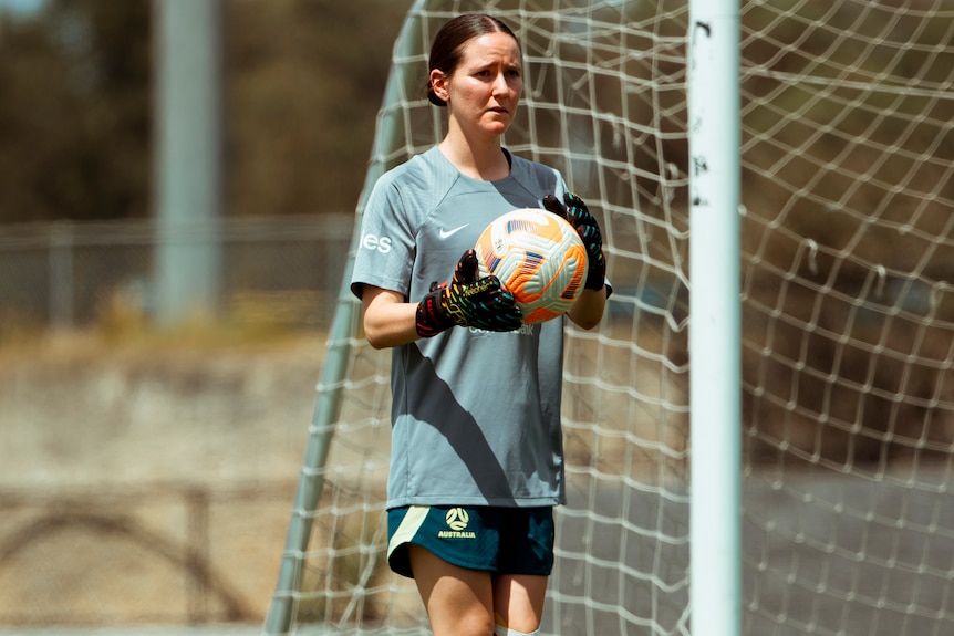 A woman in a grey soccer shirt and goalkeeper gloves holds a soccer ball in front of a soccer goal.