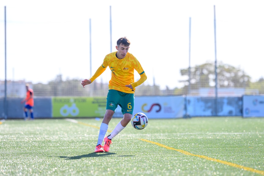 A man in a green and gold soccer uniform, white socks and pink soccer boots controls a ball during a match. He is on grass.