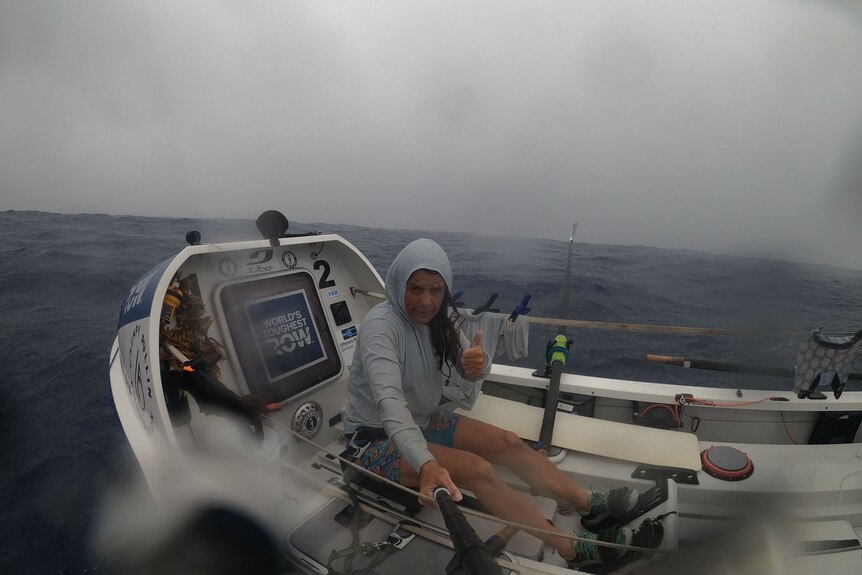 The side of a small rowboat with a woman sitting in it wearing a raincoat, with rain on the camera lens