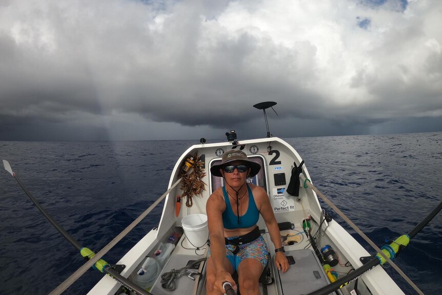 A woman seated and rowing a small boat on the ocean with storm clouds in the background