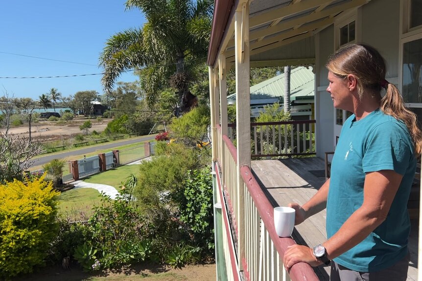 A woman standing on an upper verandah overlooking her yard out to the ocean