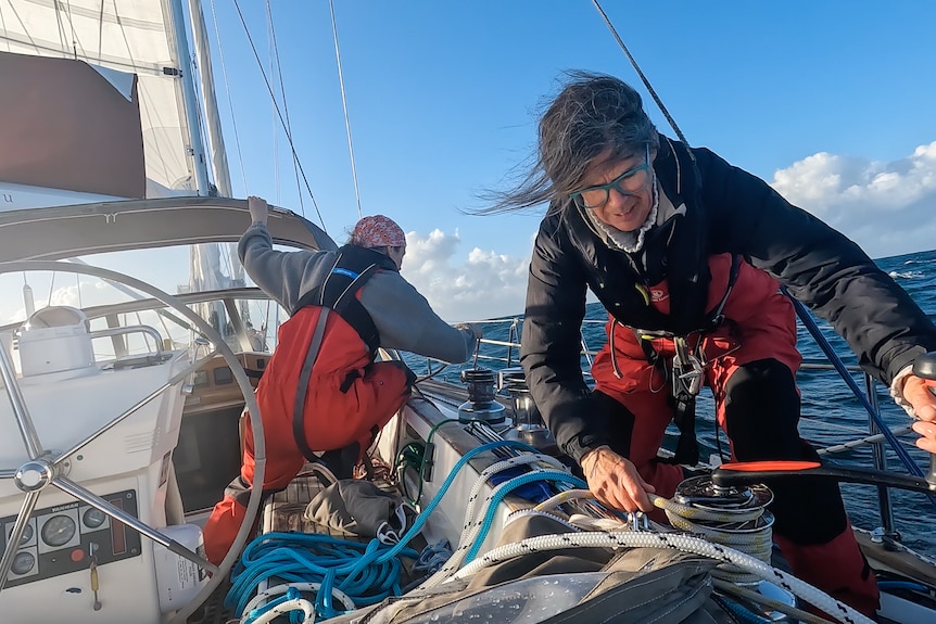 Two women are sailing on board a boat at sea - the woman in the foreground is operating a winch, the other is working with rope.