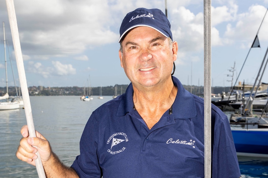 A sailor stands on the bow of his boat moored in Sydney Harbour, smiling at the camera.