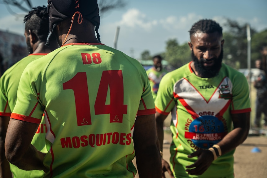 Three rugby players in uniforms walk on a dusty football field.