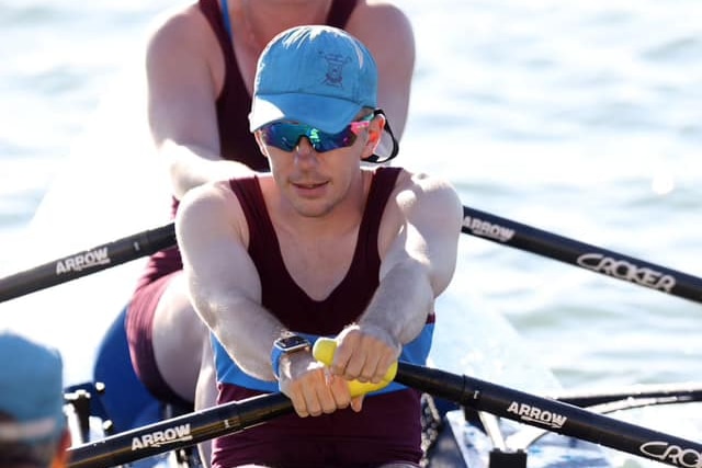 A man and a woman sit in a two person rowing boat, both wearing caps, black singlets and sunglasses. They are holding oars.