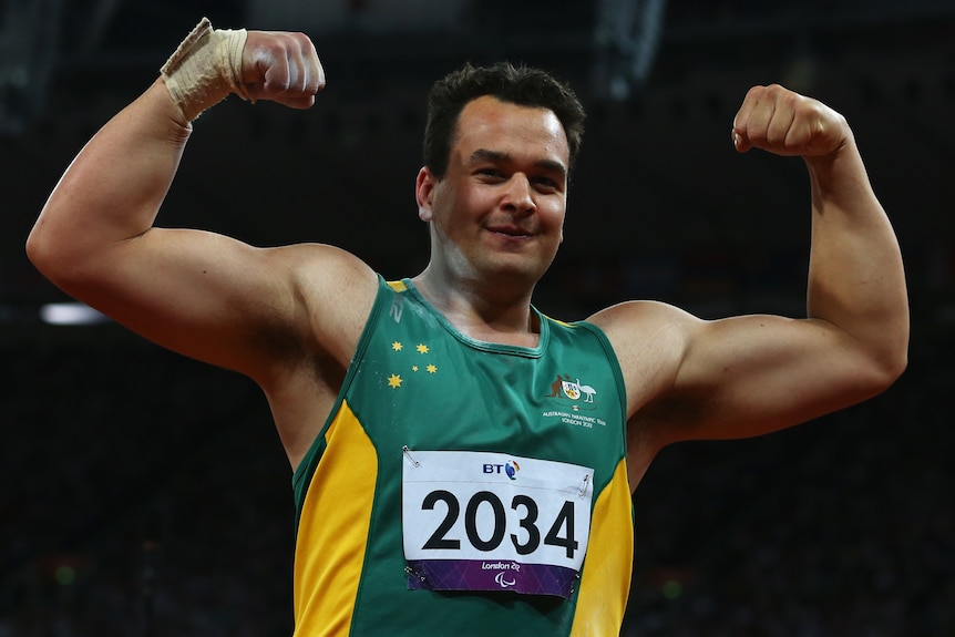 A man with short brown hair in an australian branded singlet has his arms outstretched, flexing his muscles.