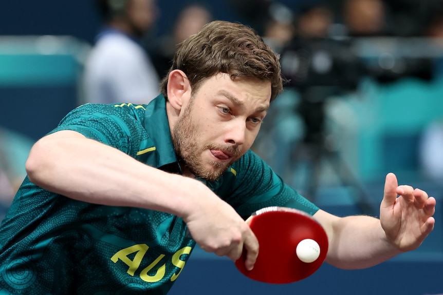 A man with short brown hair in a green Australia shirt uses a table tennis bat to hit a table tennis ball.