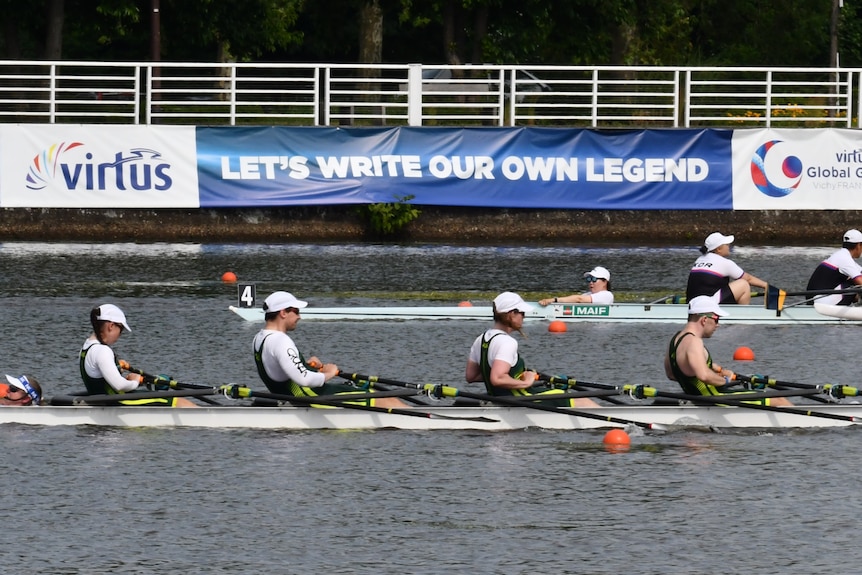 A four person rowing boat is side on from the camera. All rowers are wearing white caps and shirts.