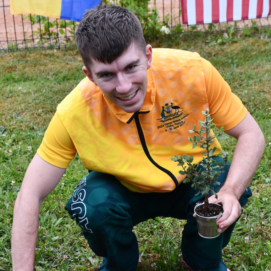 A smiling man with short brown hair in a yellow and green Australia branded tracksuit crouches and holds a small plant.