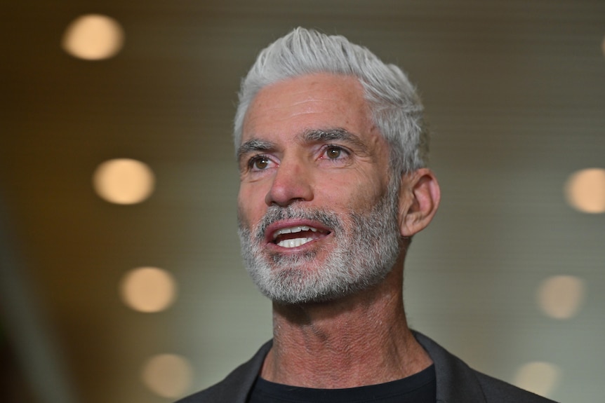 Craig Foster speaking while wearing a dark blazer and shirt in front of yellow LED roof lights