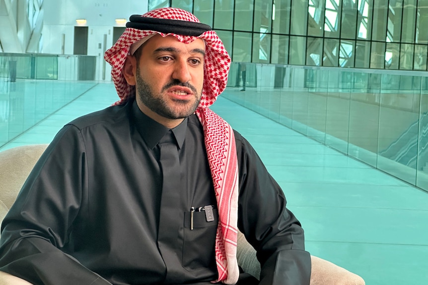 Saudi Arabia's Hammad Albalawi sitting in a cream colour chair wearing a black gown and red head covering on a blue background
