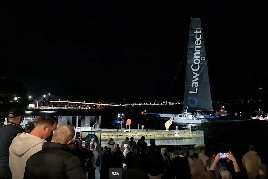 People on the shore watch on as a yacht with black sails arrives in Hobart, with the Tasman Bridge in the background.