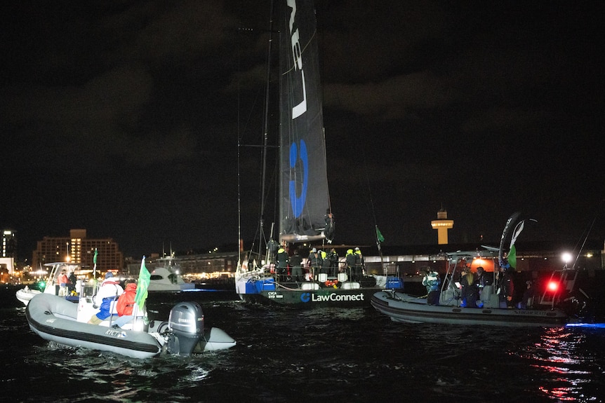 A yacht with black sails at night pulls into port at Hobart surrounded by chase boats.