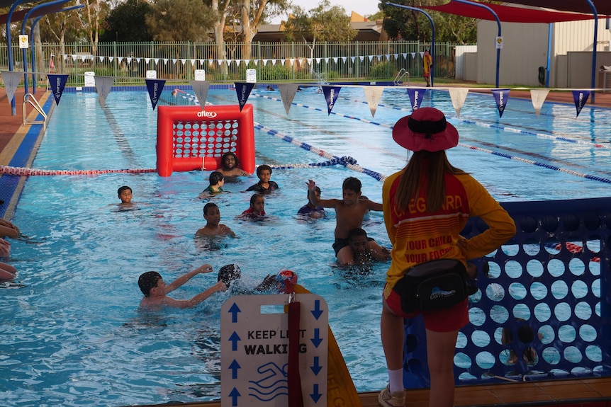 Kids playing aqua footy in a swimming pool while a lifeguard watches on.