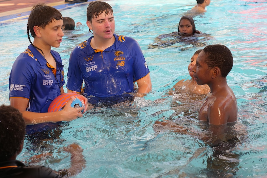 Two teenagers in blue shirt standing in a swimming pool surrounded by younger children