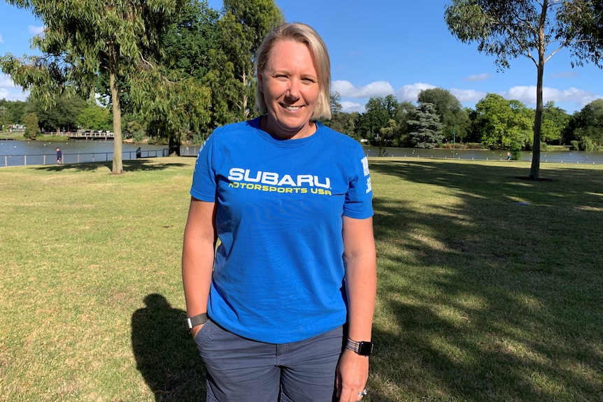 A woman with shoulder-length blonde hair wearing a blue T-shirt standing in front of parklands a lake.