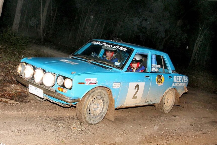 A blue Datsun rally car on a dirt road in a forest at night.