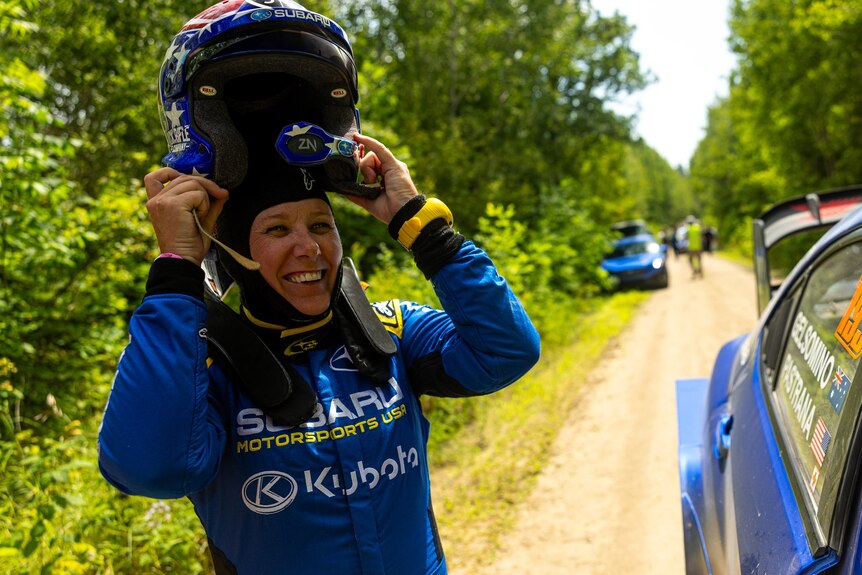 A woman wearing racing gear and removing a helmet on the side of a dirt road with rally cars in the background.