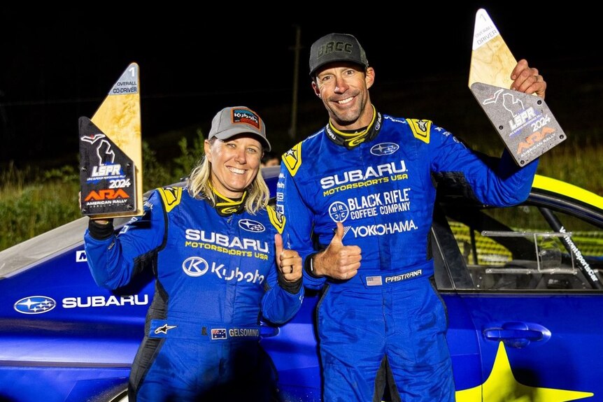 A blonde woman and taller, dark-haired man in racing uniforms standing in front of a blue car holding trophies.