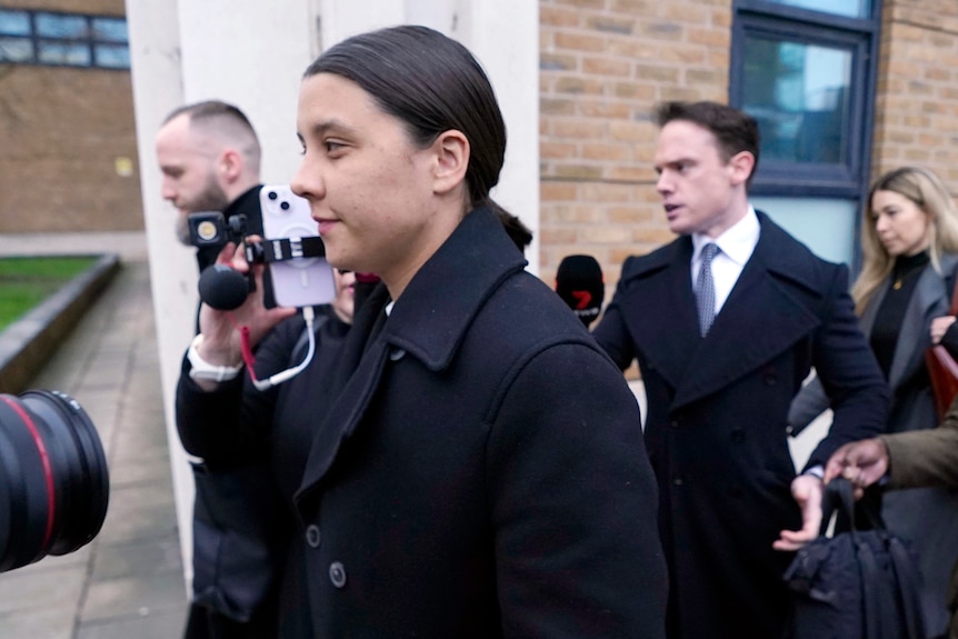 Sam in a black coat being followed by cameras and journalists. 