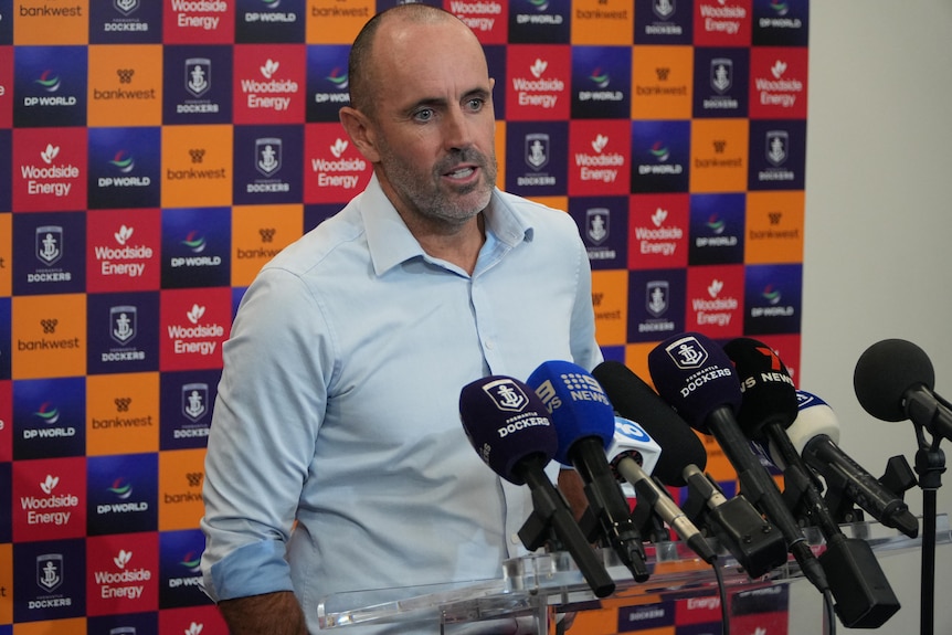A man in a collared shirt speaks in front of microphones.