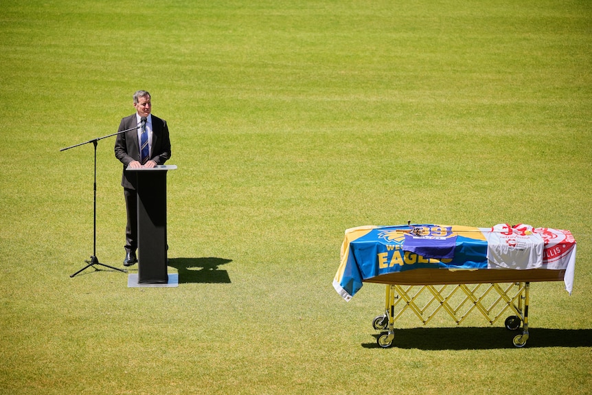 A man in a suit stands in front of a podium on a grass oval next to a coffin draped in football flags