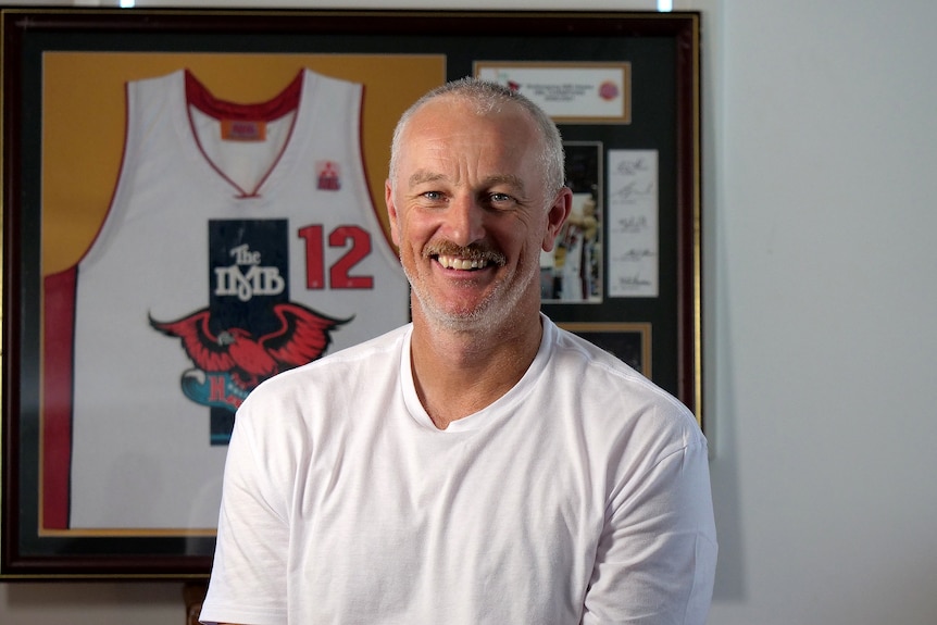 Glen Saville smiles at the camera in a white shirt with his framed Hawks jersey behind him.