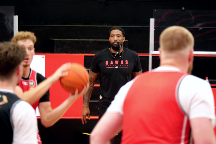 Justin Tatum wears a black Hawks shirt with a whistle in his mouth while players run in front of him.