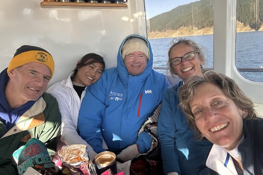 A male ocean swimmer rugged up in a jacket next to four support crew members on a boat in New Zeland