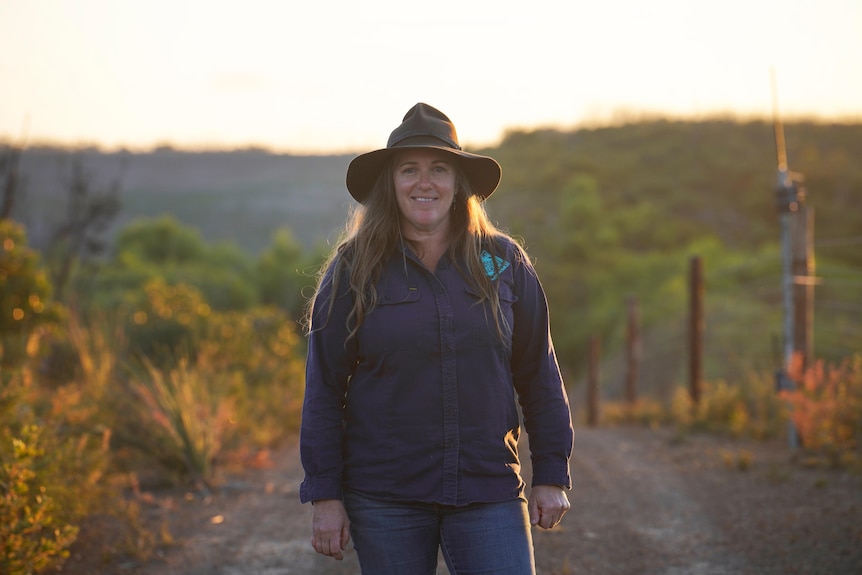 A woman in a long sleeved dark blue shirt and hat stands in a bushland area with the background out of focus at sunset