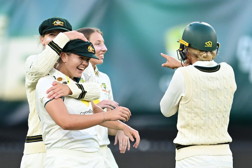 A woman cricketer smiles as she is hugged from behind by a teammate.