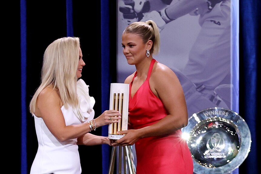 A woman in a red dress accepts a trophy from a woman in a white dress.