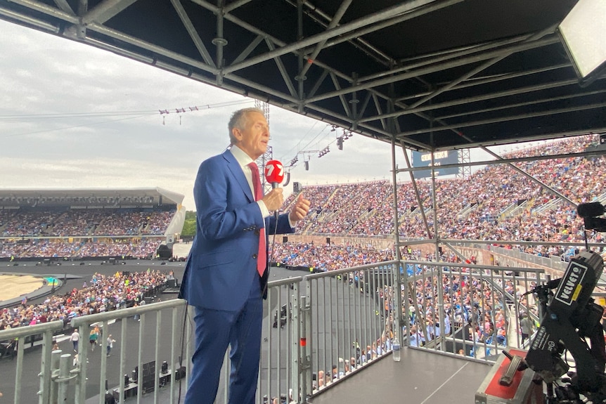 Man in suit stands on edge of packed stadium holding a Channel 7 microphone, speaking to a camera that's out of view