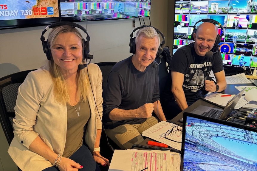 A women and two men sit at a desk surrounded by TV screens, all wearing large headphones 