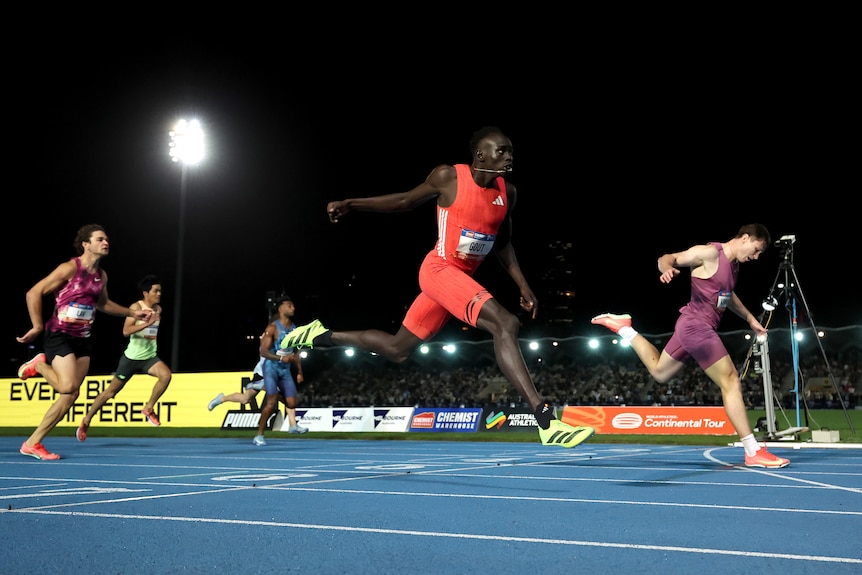 Runners cross the finish line on a blue athletics running track at night.