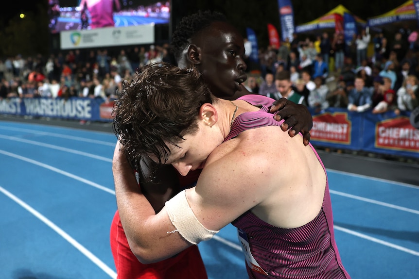 Two athletes hug on a blue athletics racing track in front of a crowd.