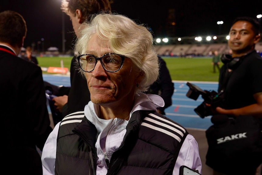 An older woman with short grey hair and black rimmed glasses stands on an athletics track.