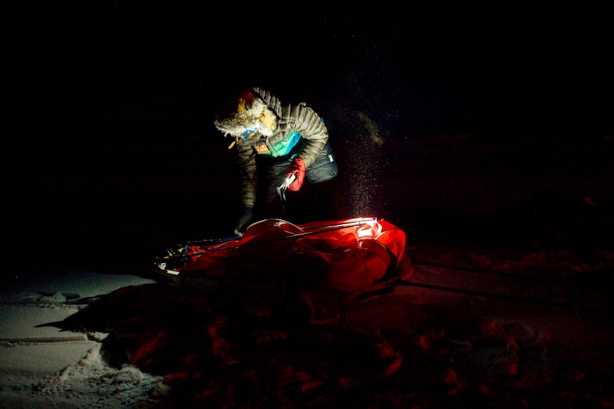 A man in warm clothing attends to a sled in the darkness.