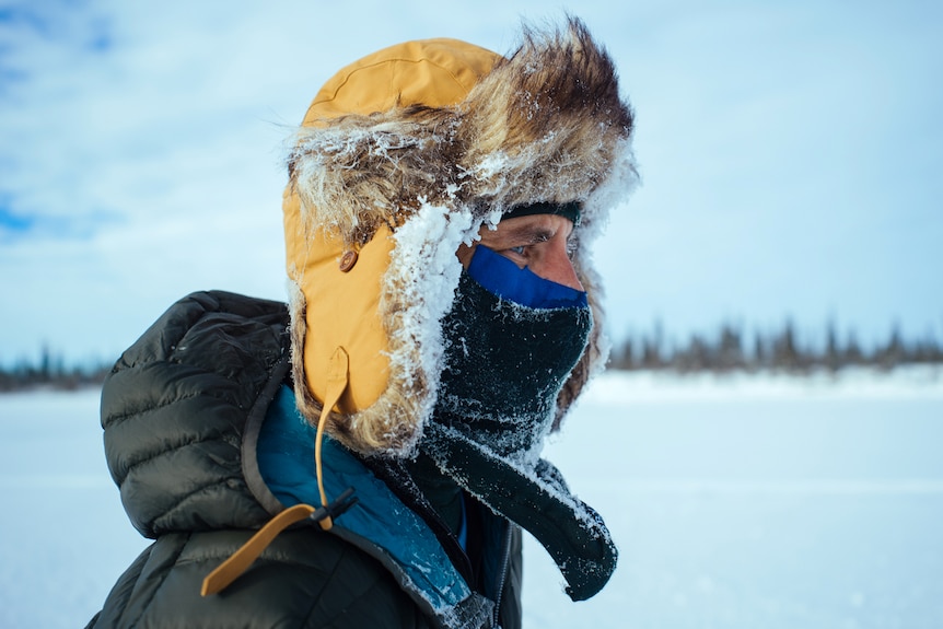 A man in a winter hat, mask and parka stares across an icy landscape.