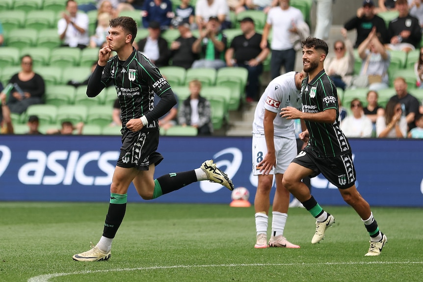 A man in a dark football shirt makes a "shushing' gesture as he runs to frame left. His teammate and a player in white in b/g