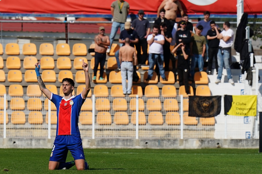 A man in a blue and red soccer jersey is on his knees with arms pointed to the sky. Crowd and yellow seats behind him.