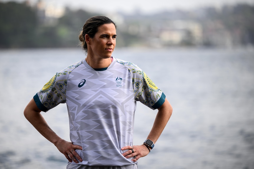 A woman wearing a white and grey soccer uniform stands with hands on hips, with a body of water behind her.