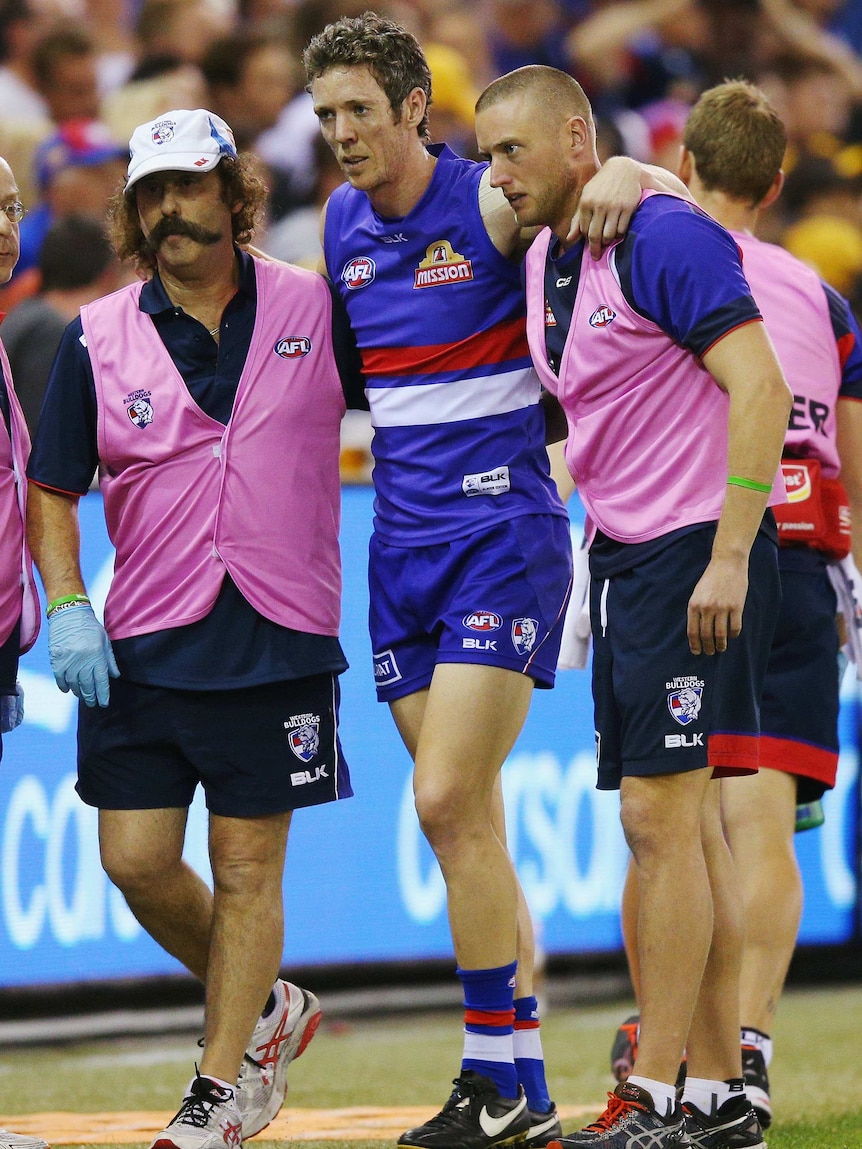 A football players supported by two men limps off the field 
